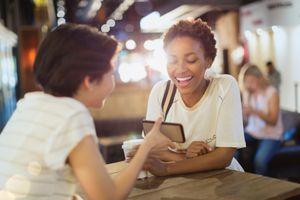 Young women friends using cell phone and drinking coffee in cafe
