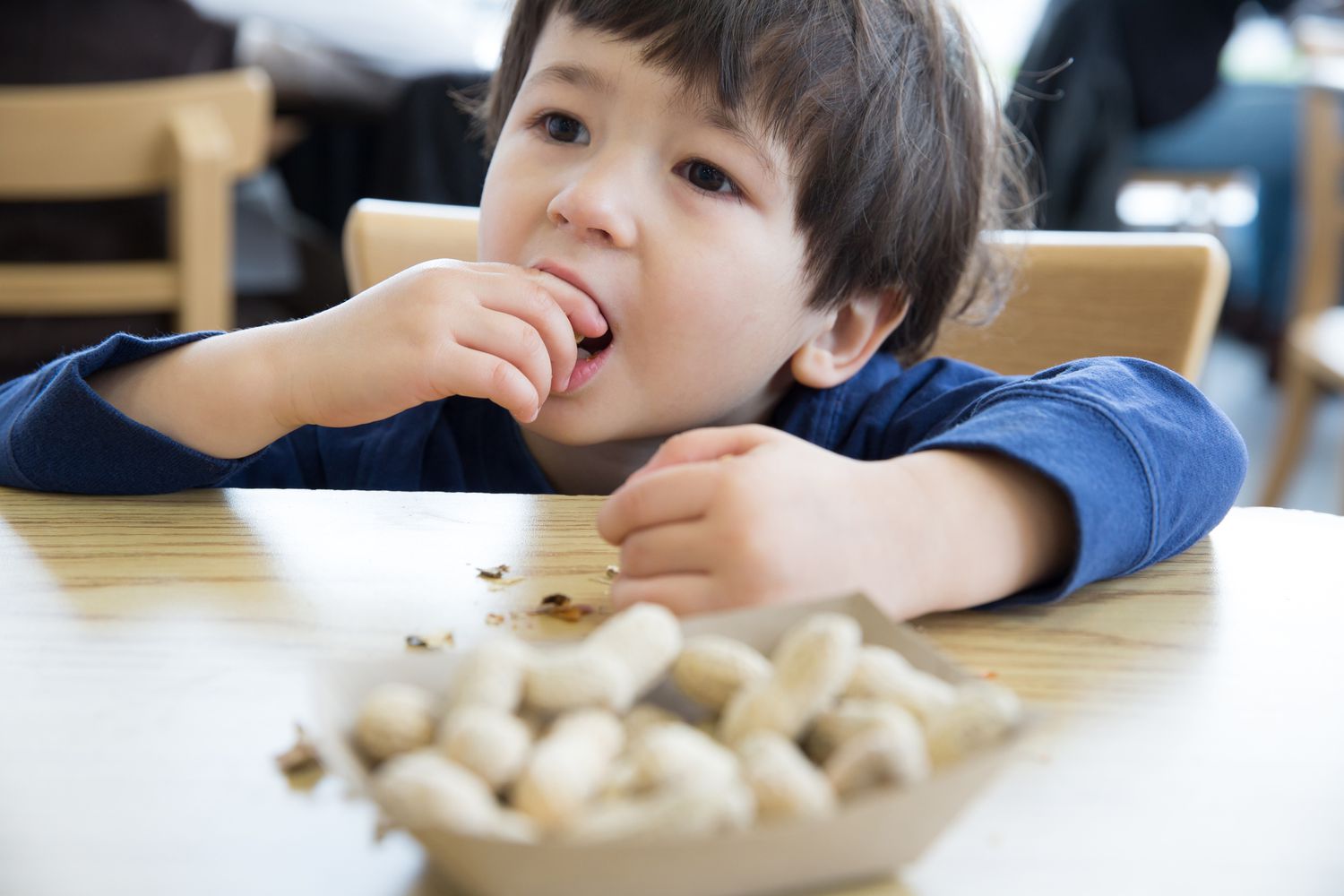 A young boy sits at a table and eats a peanut from a bowl with many peanuts.