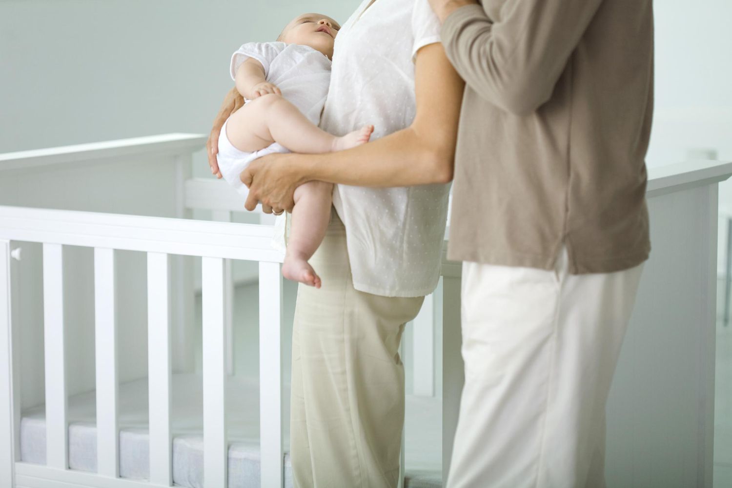 父母站在婴儿床在托儿所,妈妈lding sleeping baby, cropped view