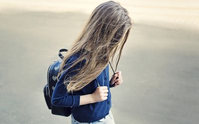 young girl with hair in her face walking outside wearing a backpack