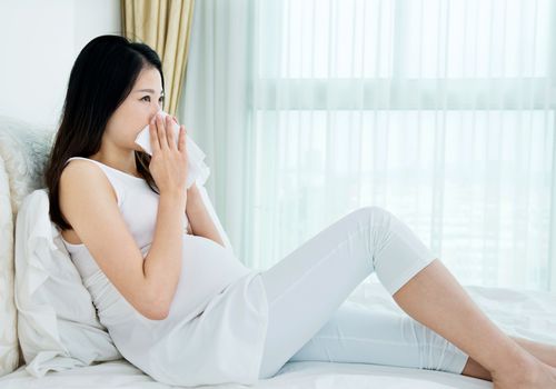 Pregnant person sitting in bed with tissue blowing nose