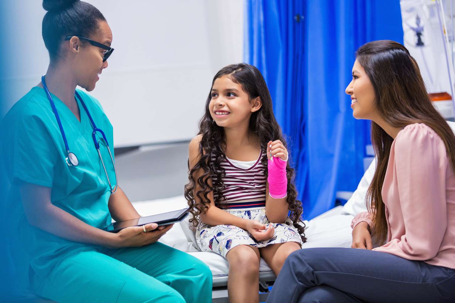a young girl with a pink cast on her arm smiling while talking to a doctor and her mother in a hospital room