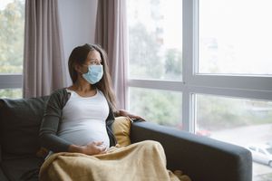 A pregnant person sits on a couch with a face mask on and a blanket while staring out the window.