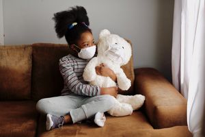 Young girl wearing a mask, holding teddy bear wearing a mask on a couch