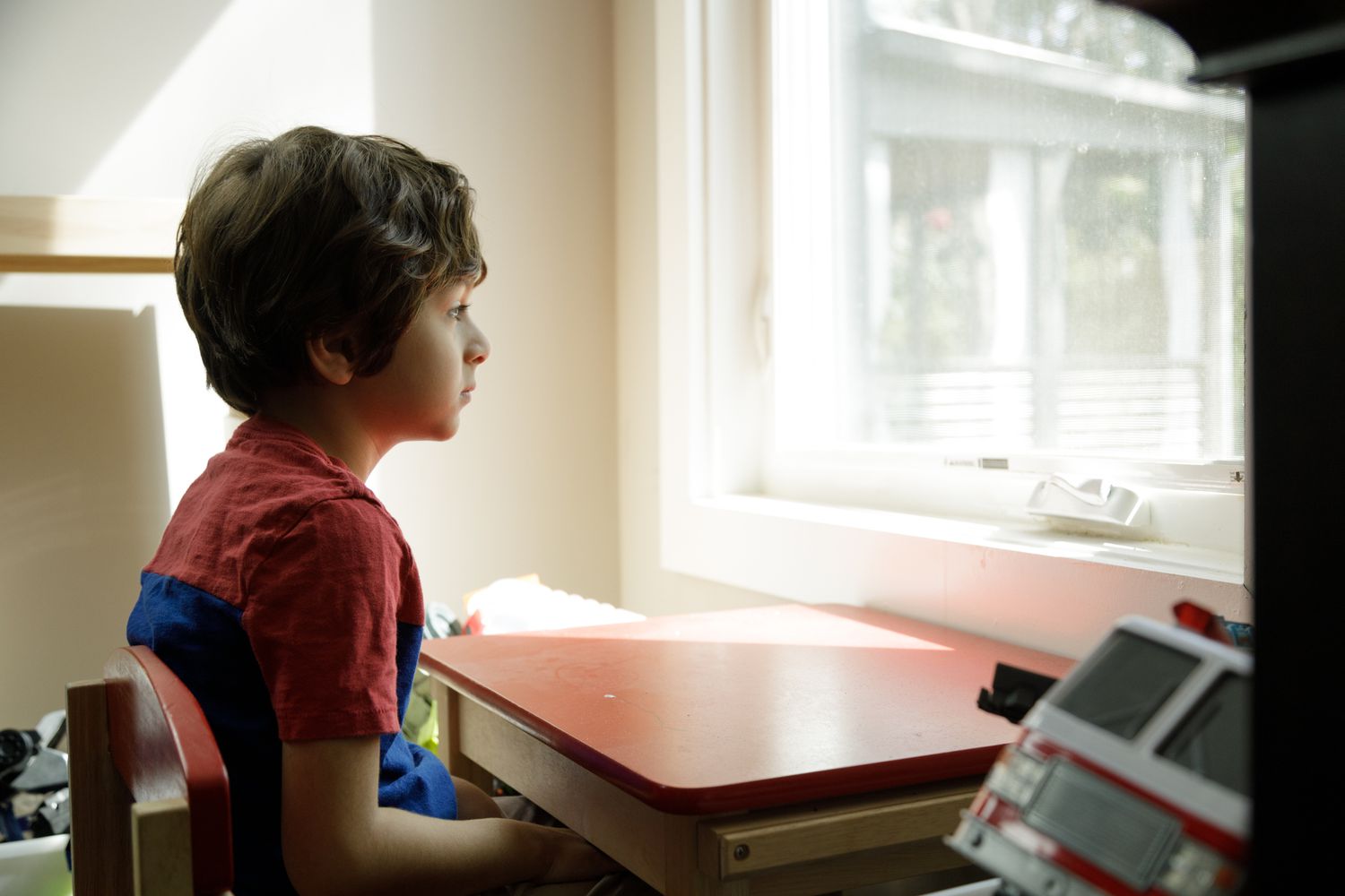 A boy with brown hair and a red shirt sits at a brown desk sadly looking out the window.