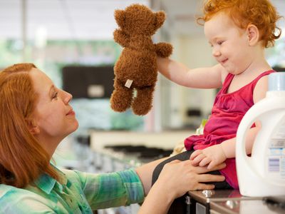 小孩抱着毛绒玩具，坐在洗衣机上面。妈妈抬头看