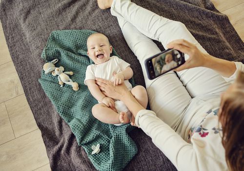 A mother takes a video of a baby with a smartphone. The baby is laying on a green blanket and is wearing a white onesie. There are toys scattered around.