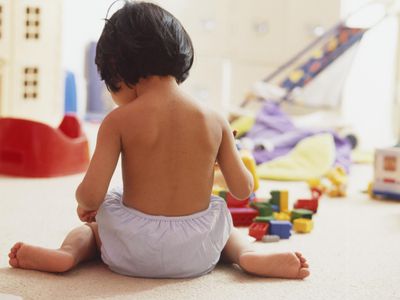 Rear view of a dark-haired girl toddler wearing trainer pants sitting on floor, surrounded by toys