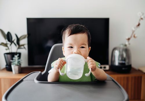 An Asian infant holds a clear sippy cup with two hands in a high chair.