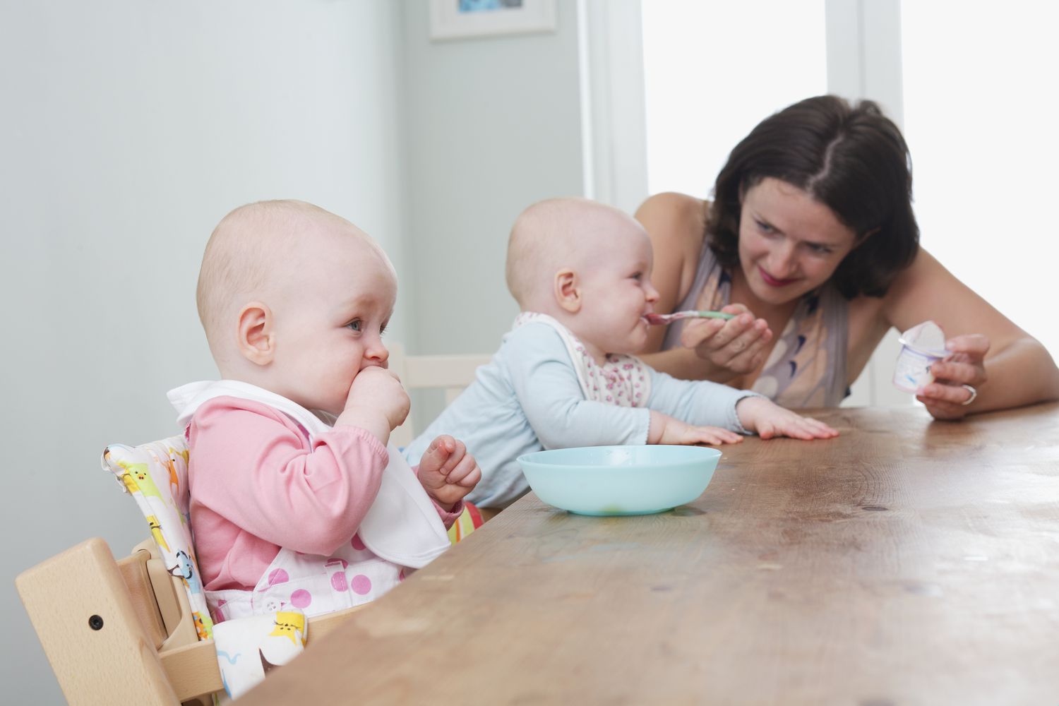 Twin baby girls seated at table, one being spoon-fed, the other girl chewing on her fist