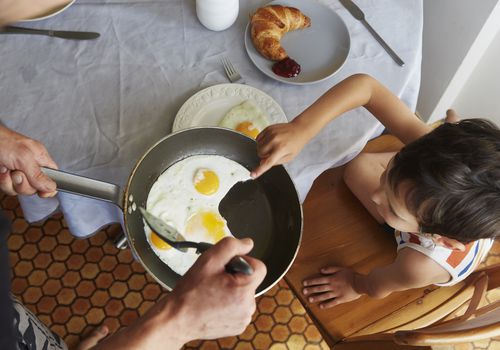 Parent offering child frying pan of sunnyside up eggs at breakfast table