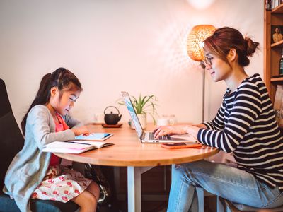 Young Asian mom working from home on the laptop computer while her little daughter reading e-learning resources on the digital tablet in the workspace at home.
