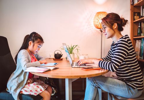 Young Asian mom working from home on the laptop computer while her little daughter reading e-learning resources on the digital tablet in the workspace at home.