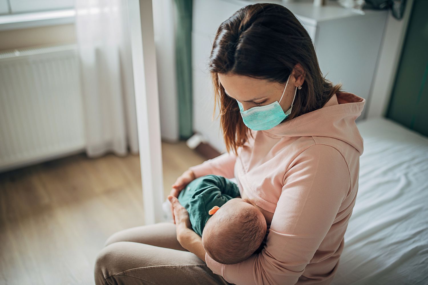 Mom wearing mask breastfeeds newborn in hospital room