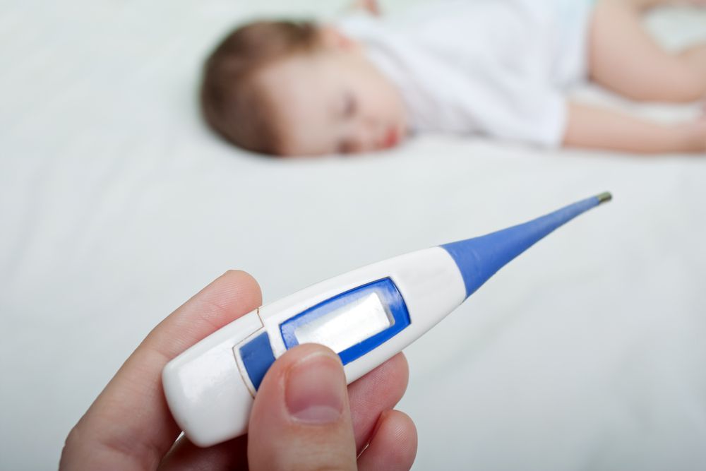 Parent holding a baby thermometer with a baby on a bed
