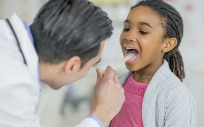Doctor applying a wooden tongue depressor to a young girl's tongue while looking in her mouth