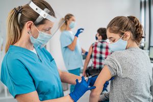 A nurse with a mask and face shield gives a teenage girl a shot.