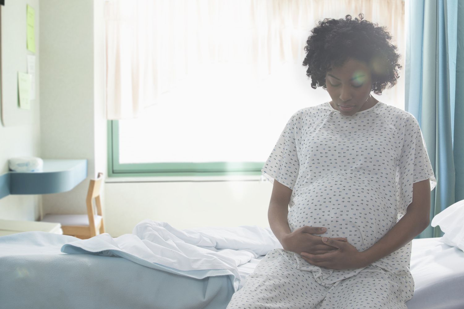 Pregnant person in hospital gown sitting on hospital bed near window