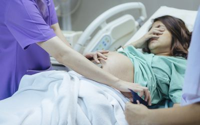 Laboring woman in hospital bed covering her face with her hand while nurse examines her.