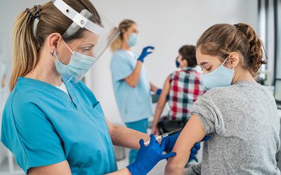 A nurse with a mask and face shield gives a teenage girl a shot.